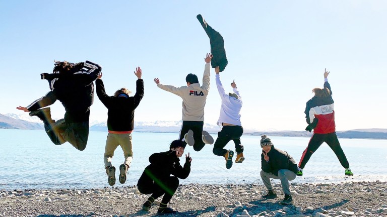 Five BTS members jump into the air while two crouch down for a picture in front of a New Zealand lake in BTS Bon Voyage Season 4